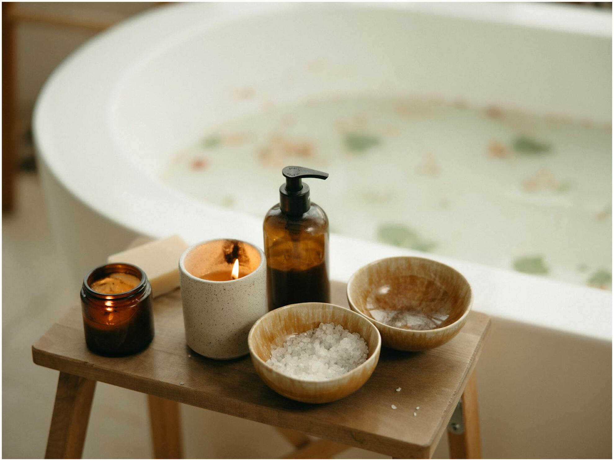 A bathtub filled with petals and milky water with a stool next to it, holding candles bath salts, and product in a bottle.