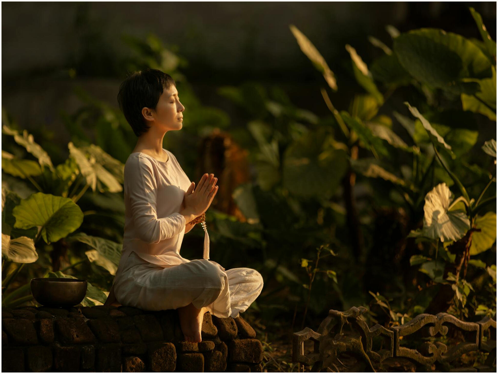 A woman in a meditative pose, sitting in a garden of lush green elephant ear plants.