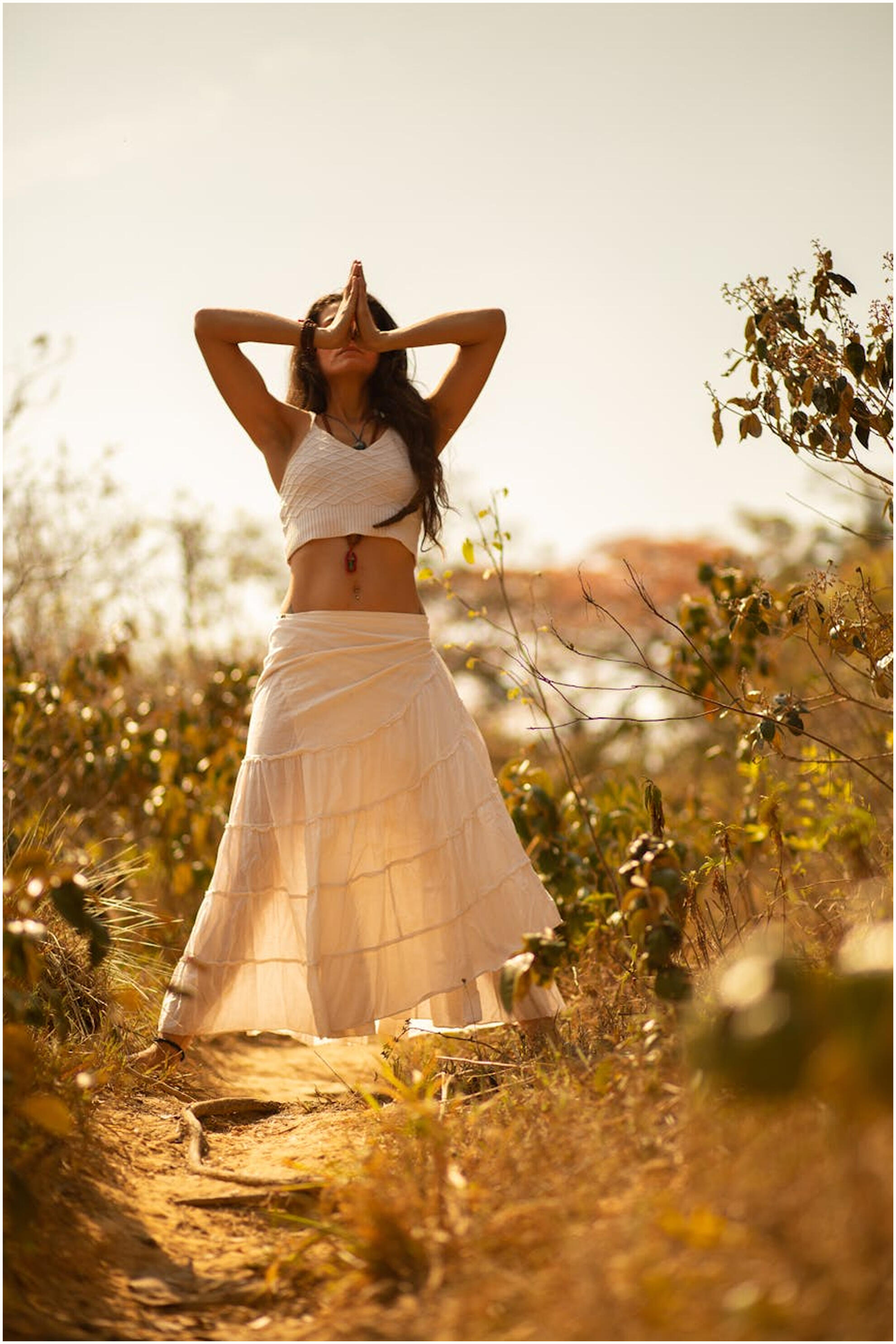 A woman standing outside in a garden with a bright sky above her. She is standing with palms together in a yoga pose and wearing a white crop top and long, flowy white skirt.