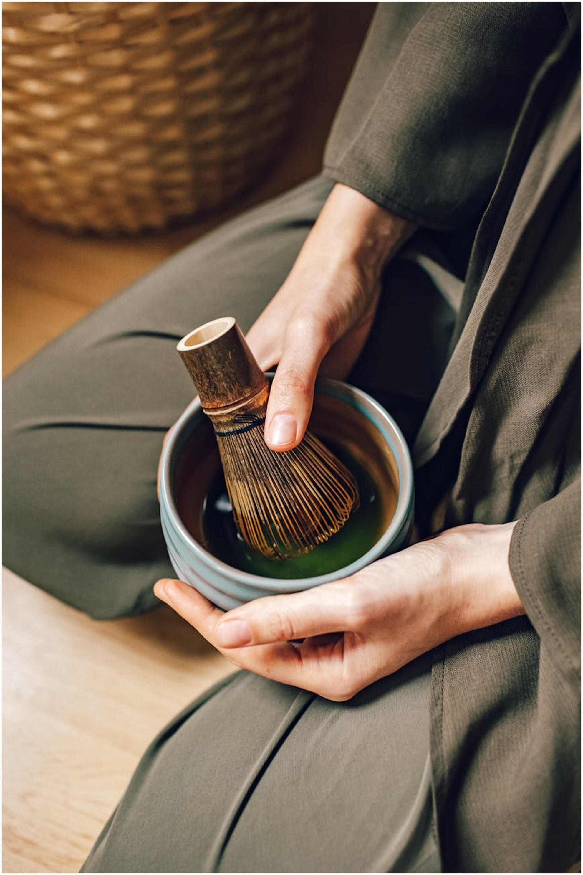 Person wearing dark green soft linens with hands holding a bowl of matcha and a matcha whisk.